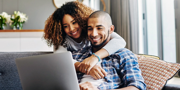 Husband And Wife Smile At Computer Screen While Learning More About Health Insurance Plans
