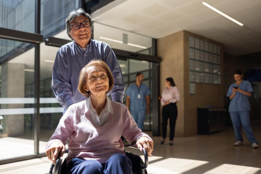 An older man pushes an older woman in a wheelchair.