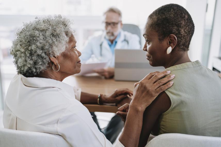 A woman with short gray hair comforts another woman with a shaved head while sitting across a table from a doctor in a white coat.