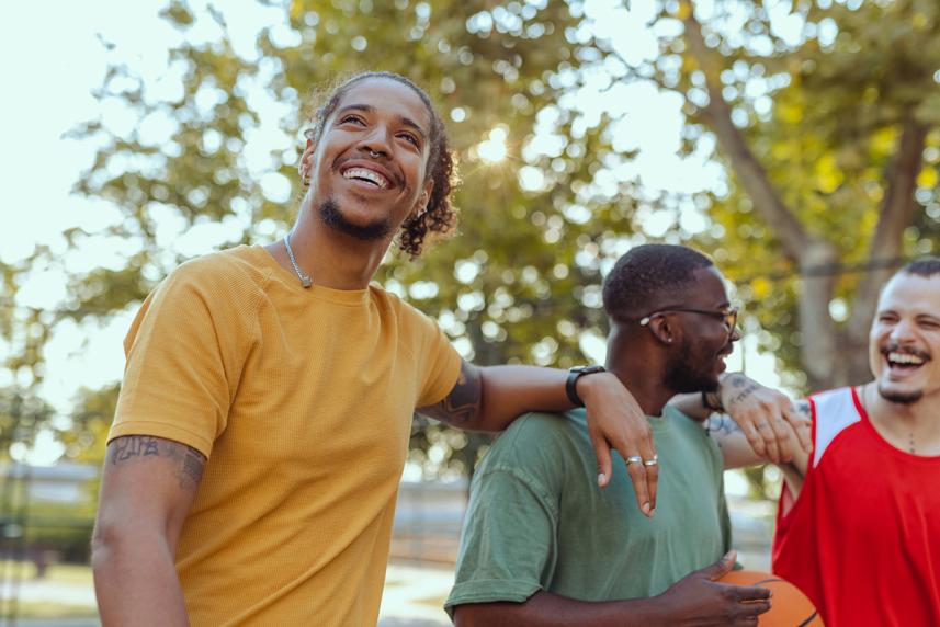 Three friends laughing together outdoors on a sunny day, one holding a basketball. They appear relaxed and happy, with trees in the background.