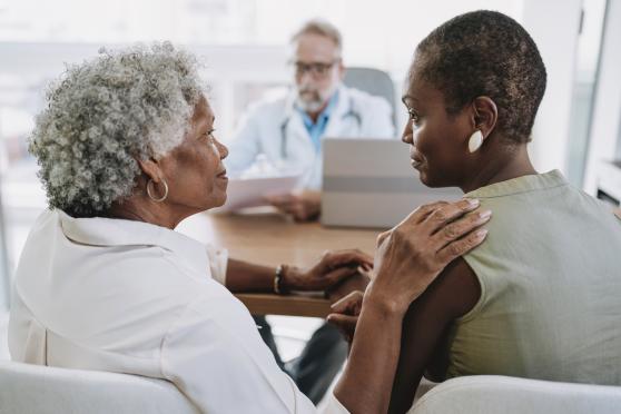 A woman with short gray hair comforts another woman with a shaved head while sitting across a table from a doctor in a white coat.