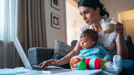 Mother On Computer Holding Child While Searching For Emotional Support Options Related To COVID-19